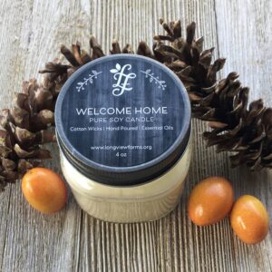 A scented candle labeled 'Welcome Home' surrounded by pine cones and acorns.